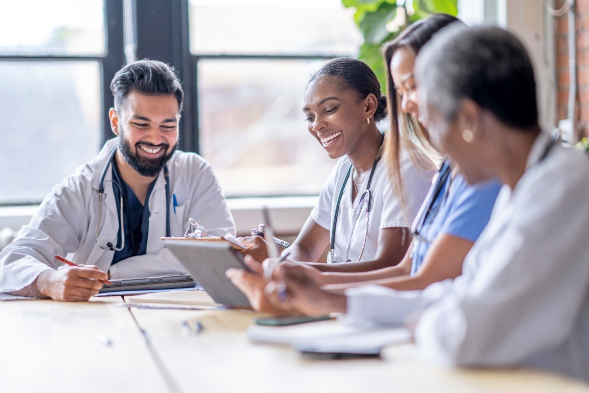 A group of four medical professionals dressed in scrubs and lab coats sit around a boardroom table as they discuss patient cases. 