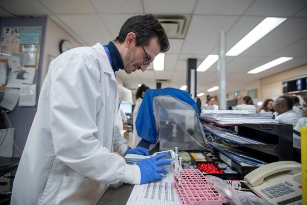 A SickKids staff member wearing a lab coat and blue gloves is working at a lab bench. In the background, other lab personnel are engaged in their tasks.